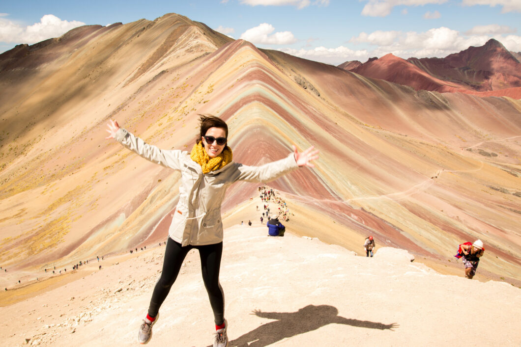 Lindsey of Have Clothes, Will Travel wearing a grey windbreaker and black leggings, jumping and smiling with Peru's Rainbow Mountain in the background.