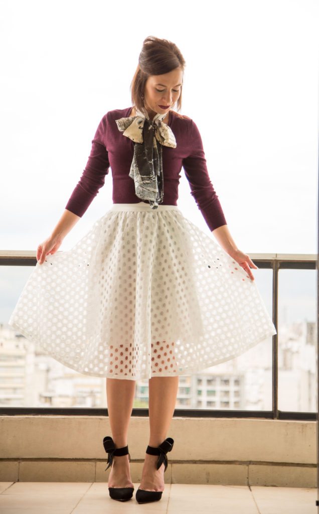 A woman poses on a scenic balcony, showing off her outfit. She's wearing a knee-length white eyelet skirt, dark maroon-colored sweater, a bowl scarf, and stiletto heels.