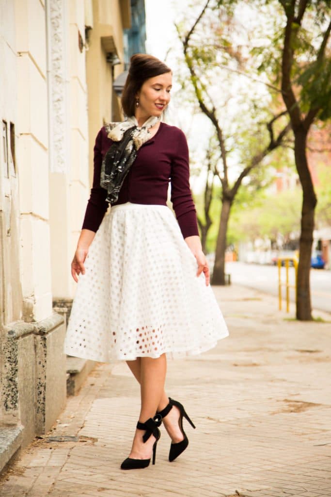 A woman poses on a city street wearing a white midi length skirt, maroon sweater, and patterned scarf. Her stiletto heels have a bow detail around the ankle.
