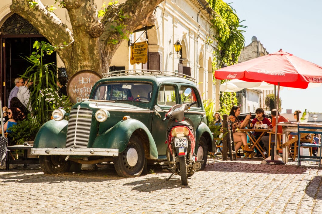 An old car and motor bike are parked outside a bustling cafe on a sunny day in Colonia del Sacramento, Uruguay