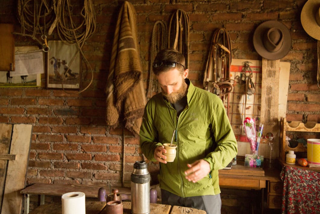 A man makes a drink while standing in a hiking cabin. He's wearing a green pull-over jacket and gray hiking pants.