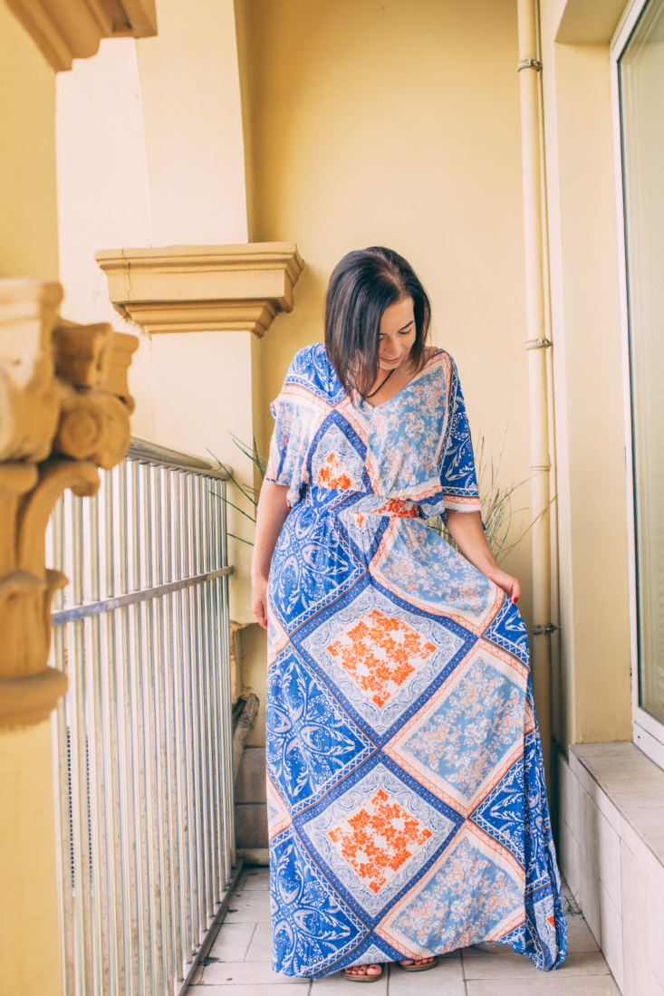 A woman stands on a hotel room balcony, admiring the floral print on her floor-length maxi dress. The dress pattern has shades of blue, orange, and peach throughout the design.