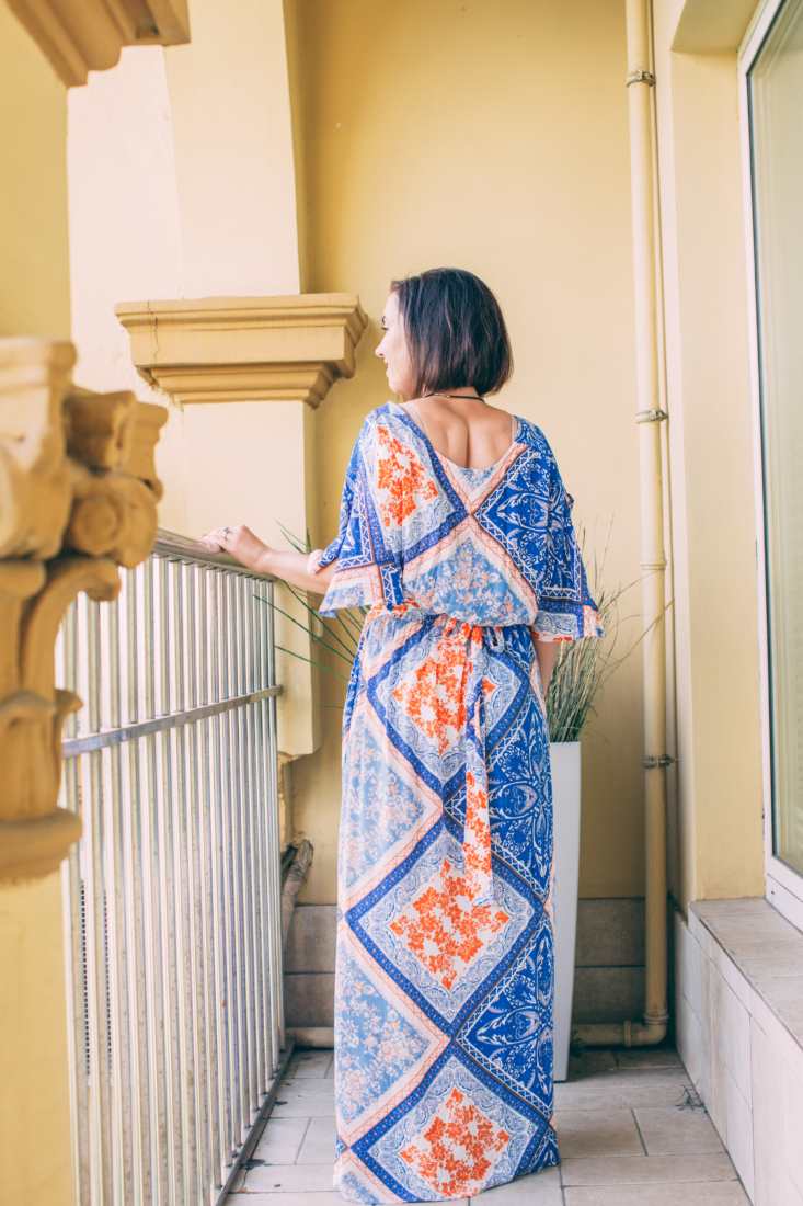 A woman poses on a hotel balcony with her back to the camera. She has short brown hair and is wearing a blue and orange floral patterned maxi dress.