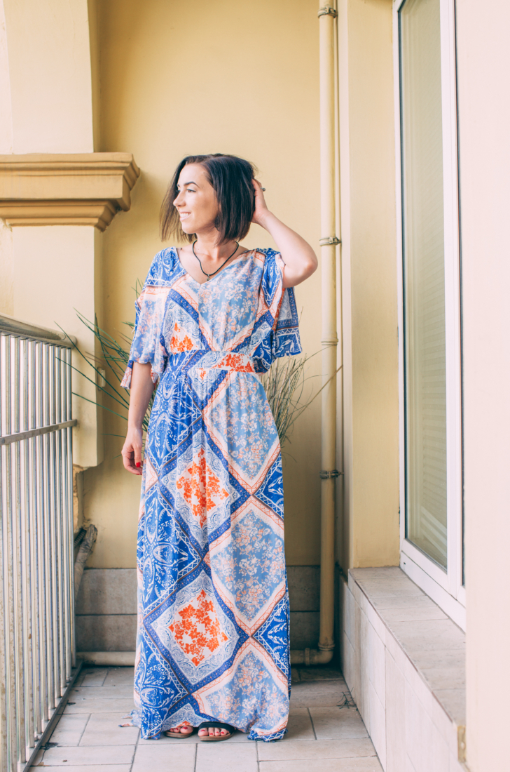 A young woman with short brown hair poses on a cement balcony, looking out over the scenery of Egypt. She's wearing a floor-length dress with a light and dark blue and orange vintage floral pattern across the dress.