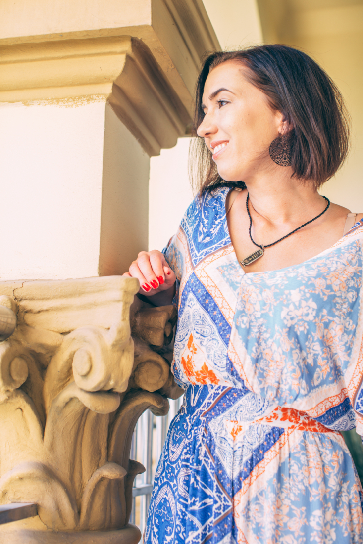 A woman poses on a hotel room balcony, looking out over the landscape. She is wearing a blue and orange floral print dress, necklace, and earrings.