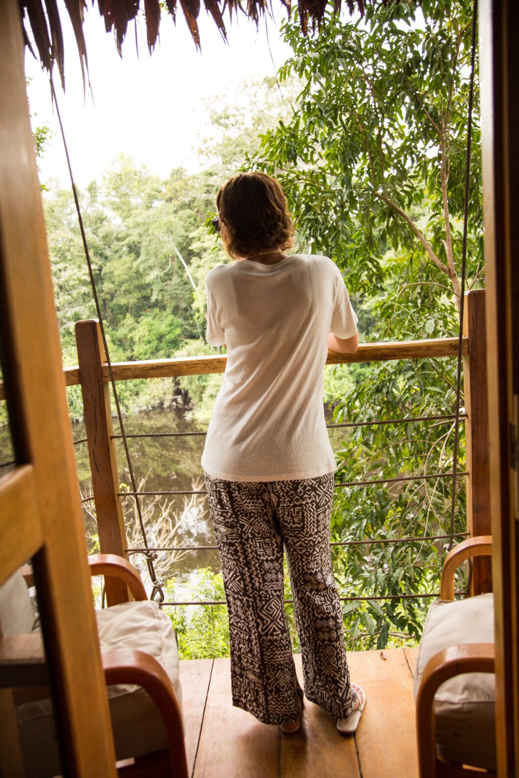 A woman stands at a balcony, looking out across the tropical forest landscape. She has her back turned to the camera and is wearing a white shirt and black and white patterned pants.