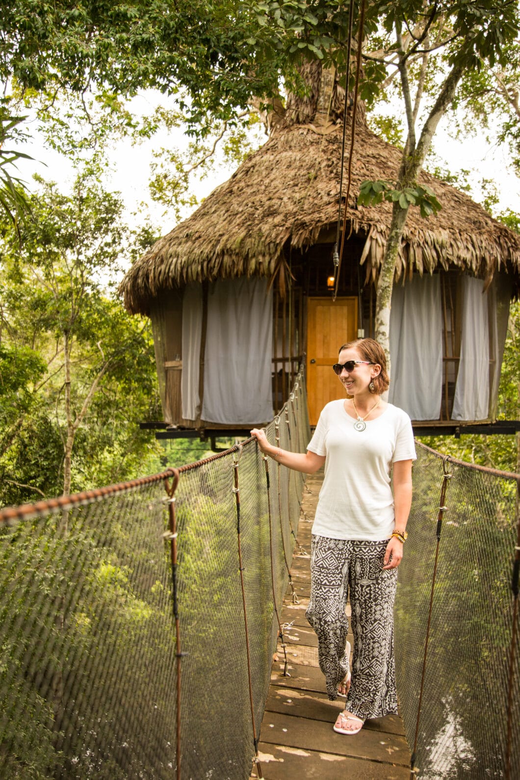 A woman walks along a wooden footbridge connected to a hut surrounded by tropical forest. She's wearing a white short-sleeve shirt and flowy blank and white patterned pants and matching accessories.