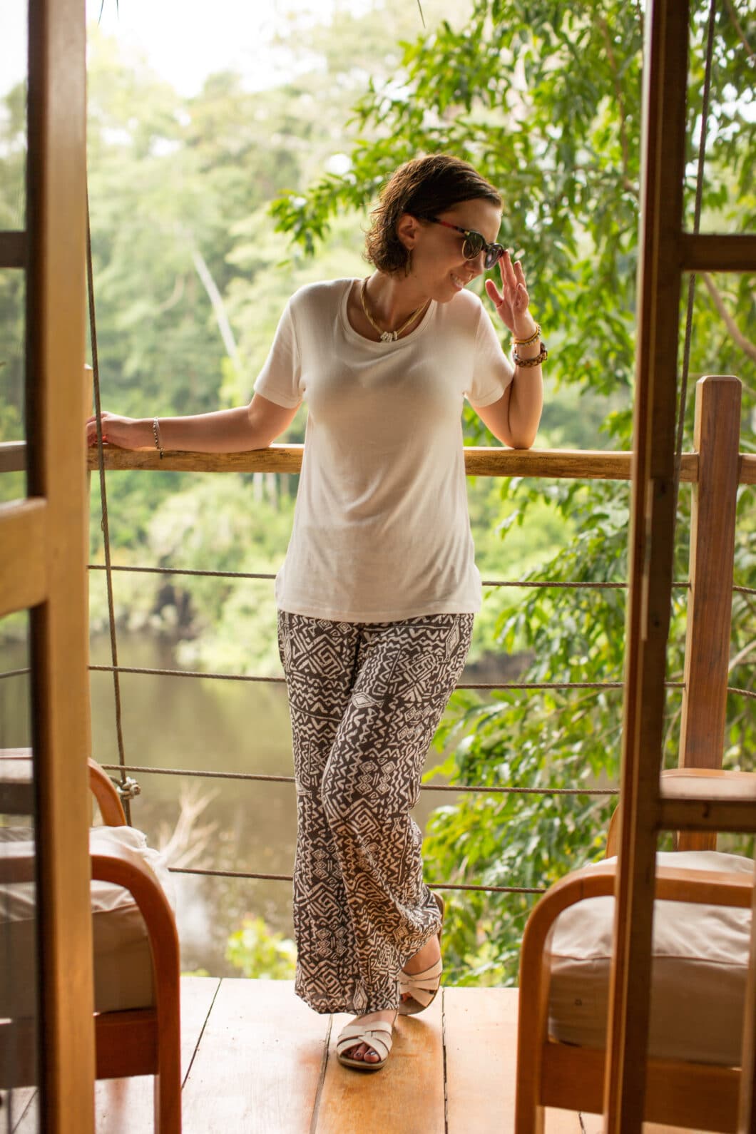 A woman poses in a comfortable, casual black and white outfit, standing on a balcony in front of a tropical landscape of the Amazon rainforest.