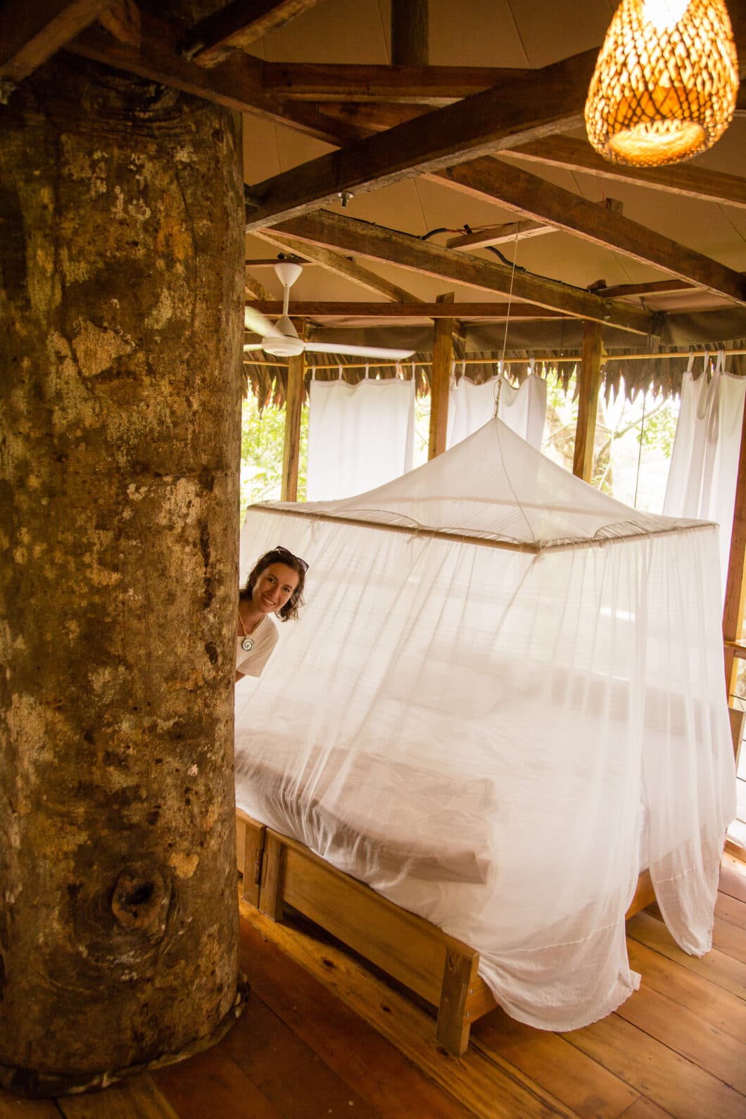 A woman peaks out from around a giant tree trunk in the center of a luxury hut. Behind her, a large bed is canopied with white netting.