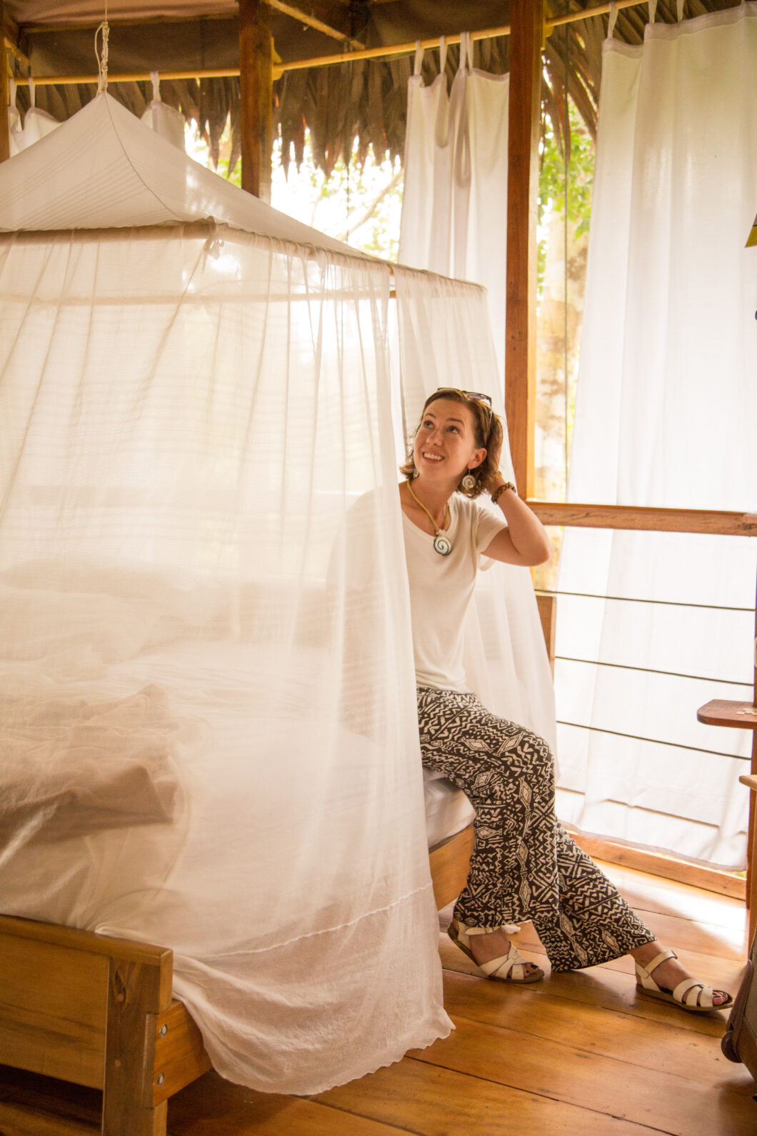 A woman sits on a bed inside a luxurious tropical hut. The bed is canopied in white mosquito netting.