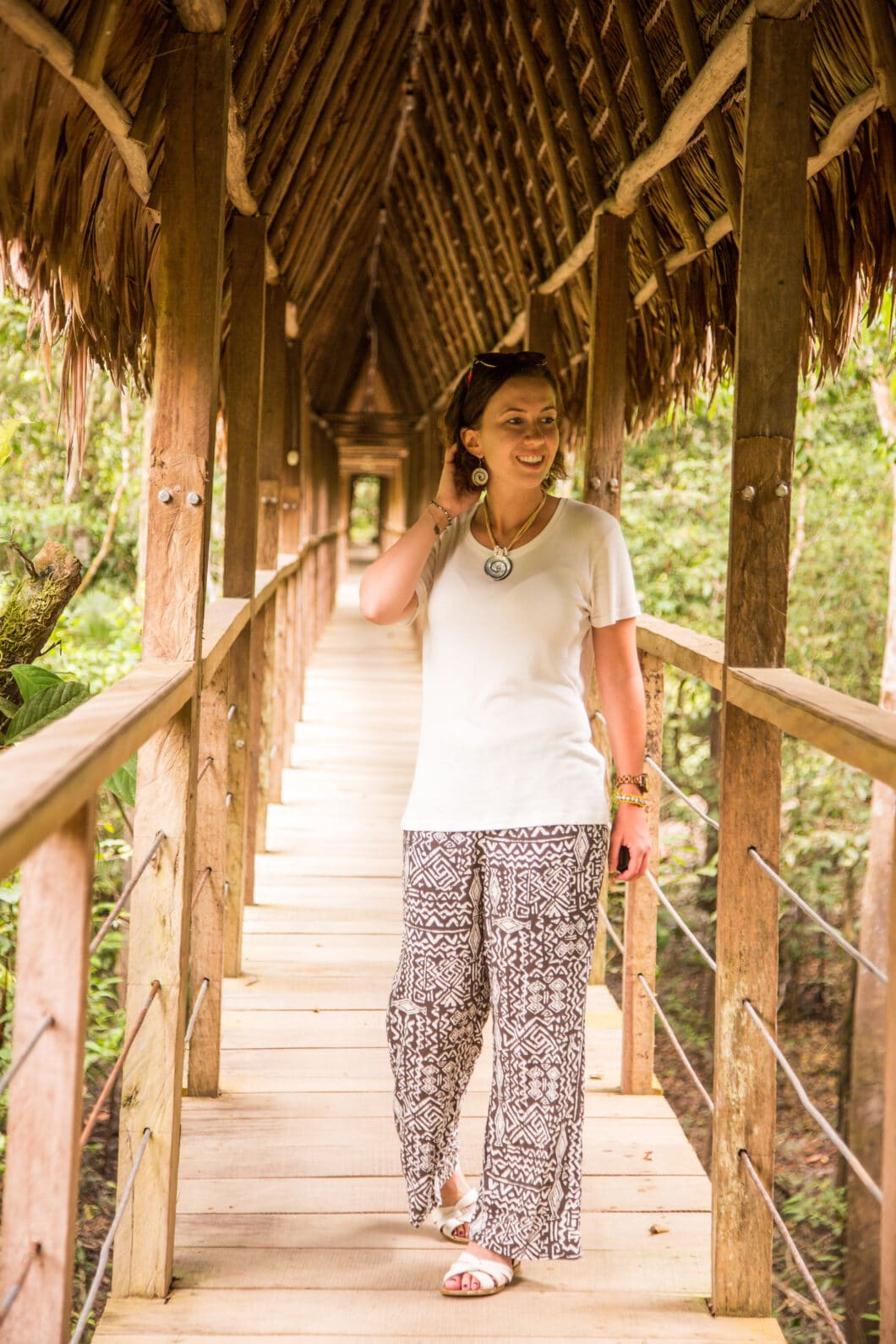 A woman poses on a covered wooden bridge in the Amazon Rainforest. She's wearing a casual, lightweight outfit for heat and humidity, including a white t-shirt and flowy black and white pants, paired with casual accessories.