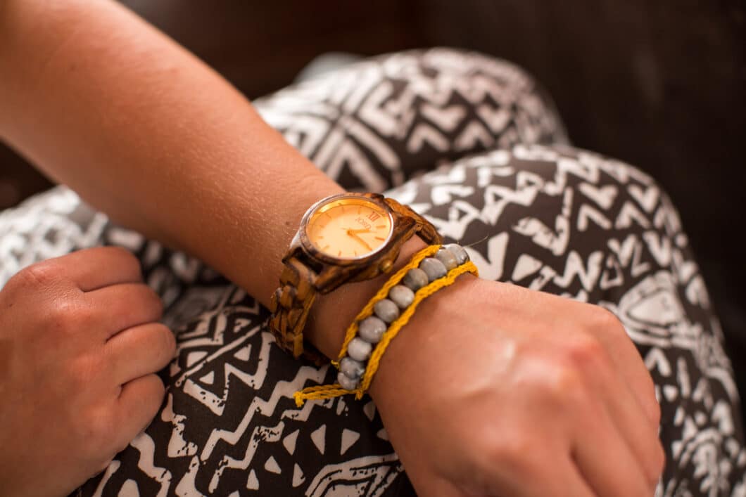 A woman's arm is pictured, showing a wristwatch and beaded bracelet on her wrist. Her arms rests across her legs, and she's wearing black and white patterned pants.