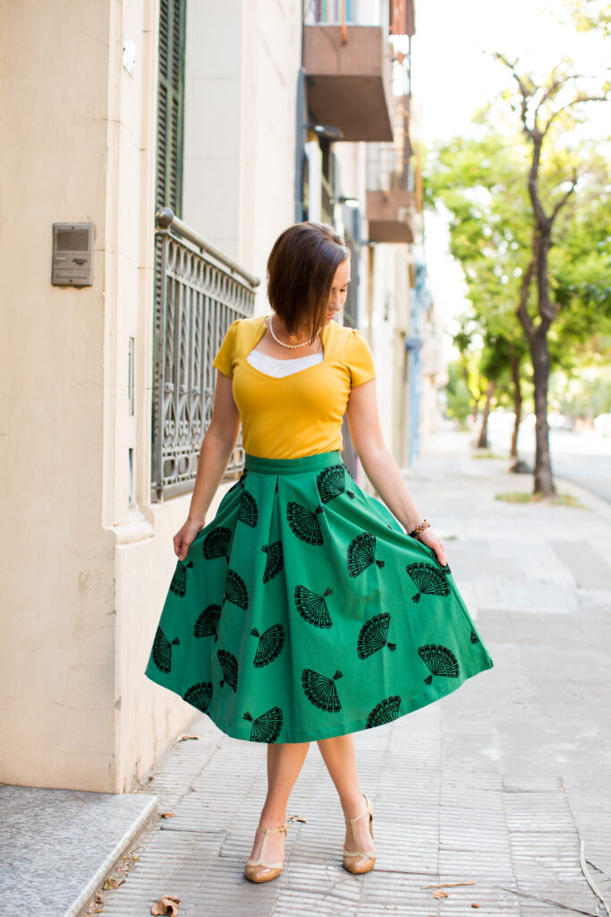 A woman poses on a city street wearing a flared patterned green skirt and yellow top, paired with nude t-strap heels.