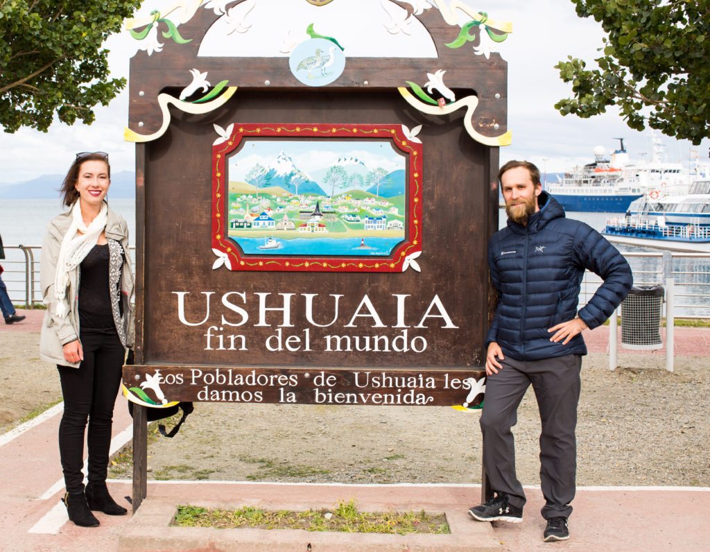 A man and woman posing in front of a board in Ushuaia Argentina that reads 'Fin del Mundo' in Spanish.