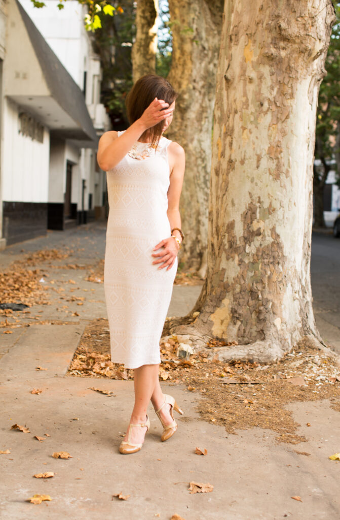 A woman poses near a large tree along a city street. She's wearing a below-the-knee white sleeveless dress and nude t-strap heels.