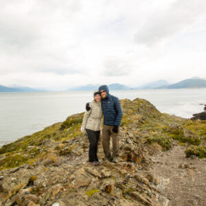 Zac and Lindsey of Have Clothes, Will Travel bundled up in warm clothing standing on Bridges Island, Ushuaia with the sea in the background