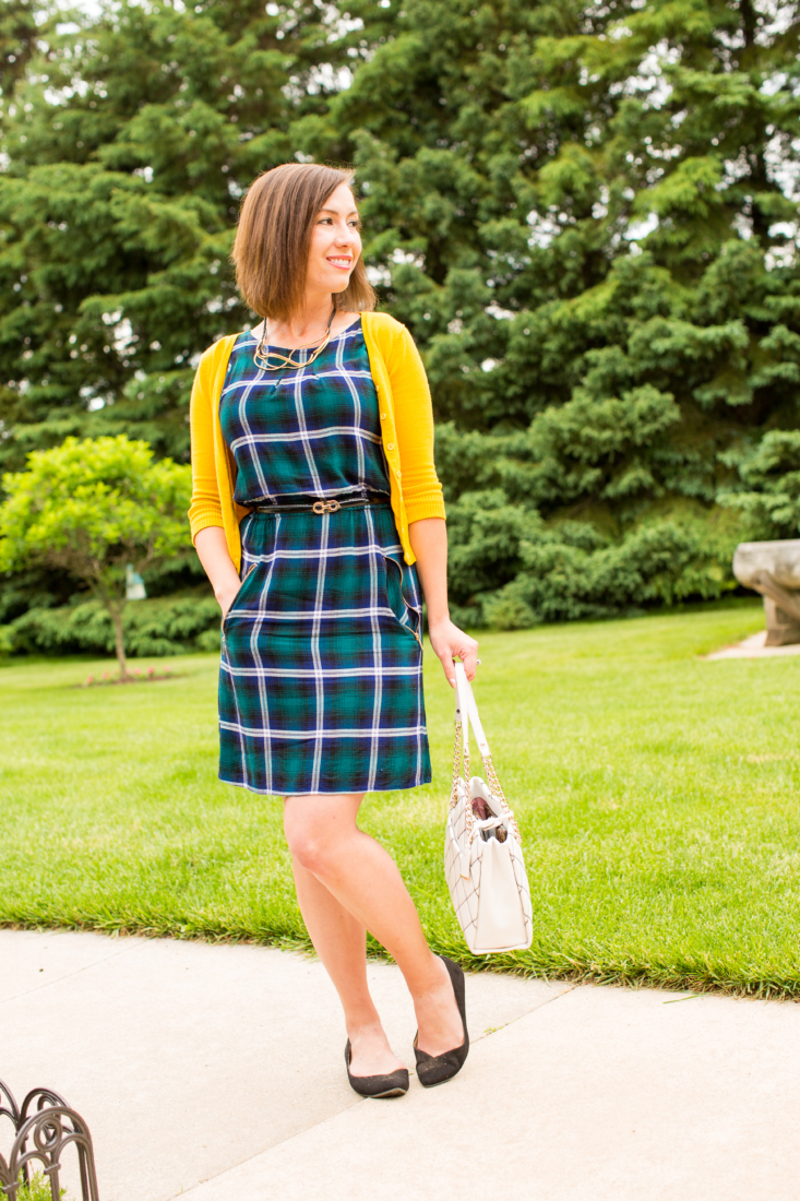 Lindsey poses outdoors in a park. She's wearing a green and white plaid dress, a yellow cardigan, and holding a white handbag.