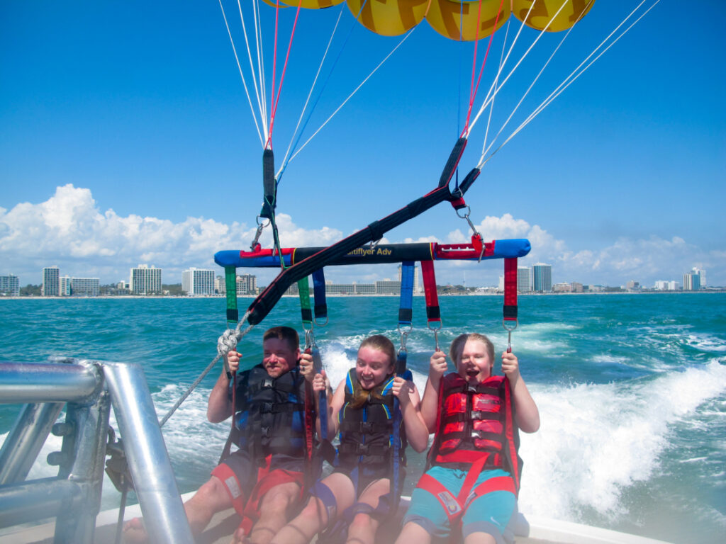 Three people are wearing harnesses and life vests on the back of a boat about to launch a parasail. The people look excited and nervous as they're about to fly over the aqua-blue water. 