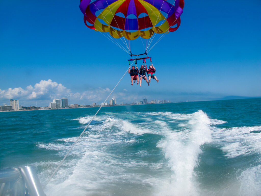 Three people are strapped into a parasailing harness with an open blue, yellow, and red parasail above them. They are riding several feet above the water, with the city skyline in the background. 