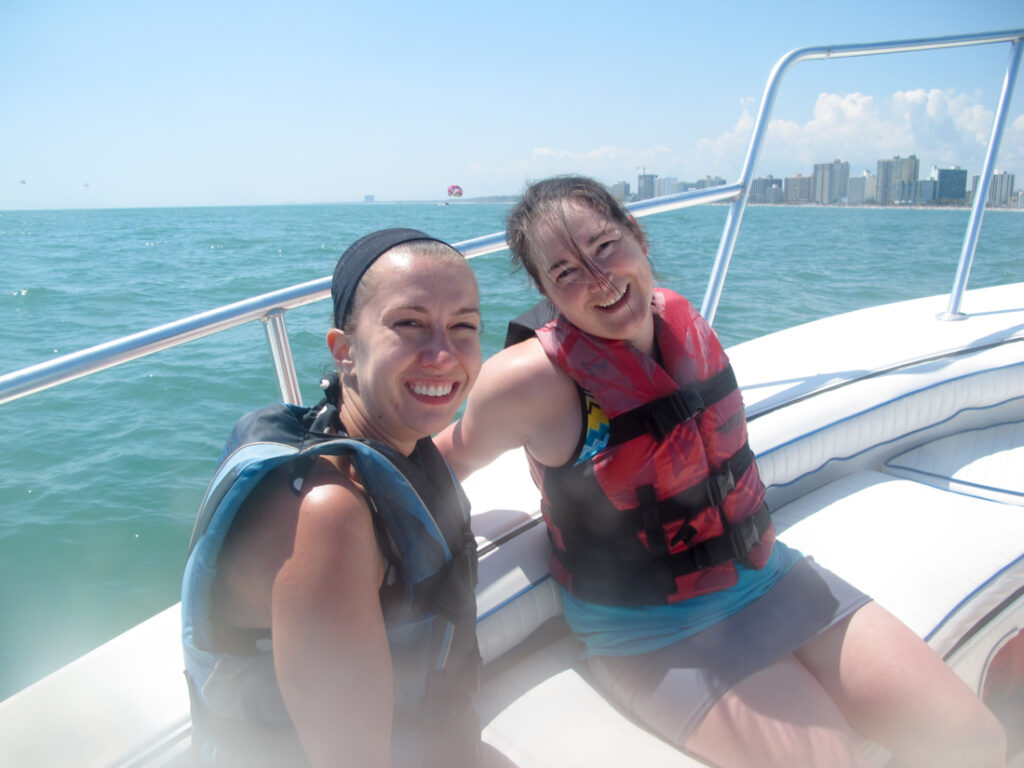 Two people are wearing life vests and bathing suits on a boat. They are smiling as they lean against the silver railing of the white motorboat with a city skyline in the background. 