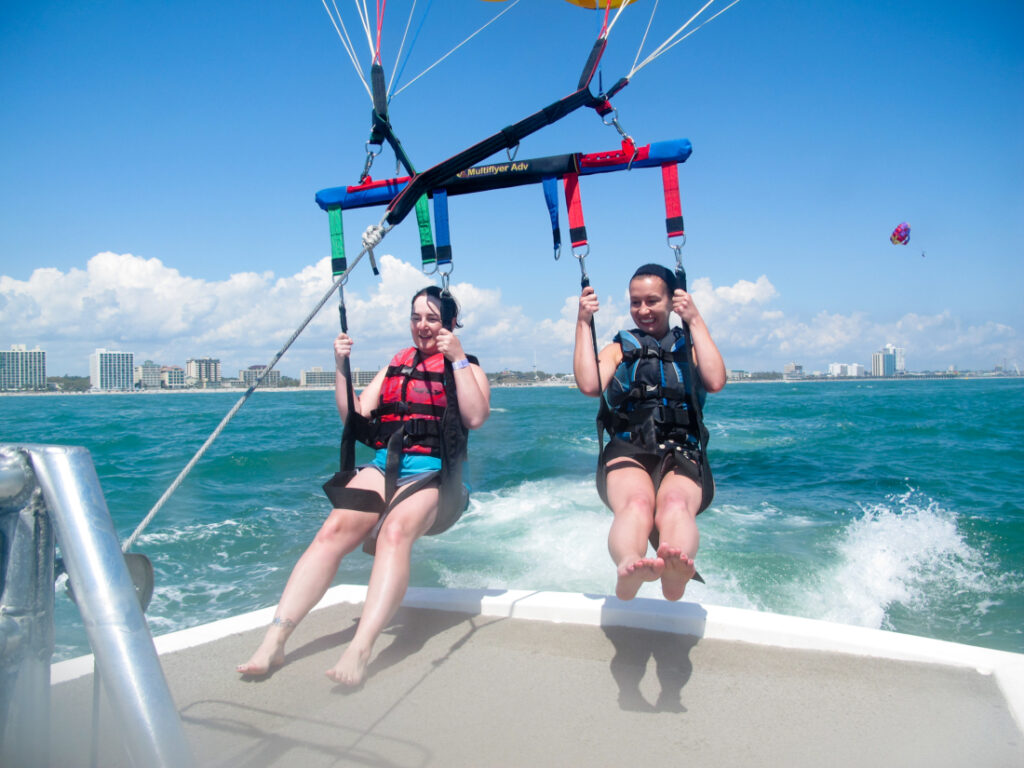 Two parasailing riders are strapped into a harness, waiting to launch off the back of a motorboat.