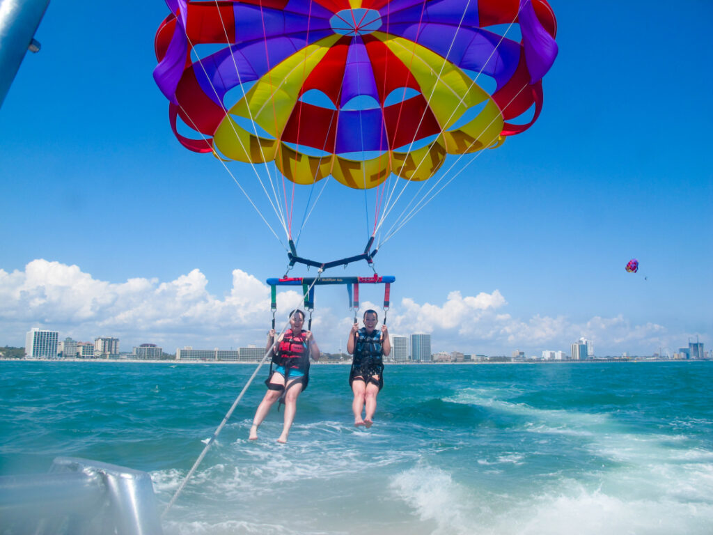 Two people with their legs extended are landing in their parasailing harness. The red, yellow, and blue parasail is open behind them, and their feet are touching the aqua-blue water.
