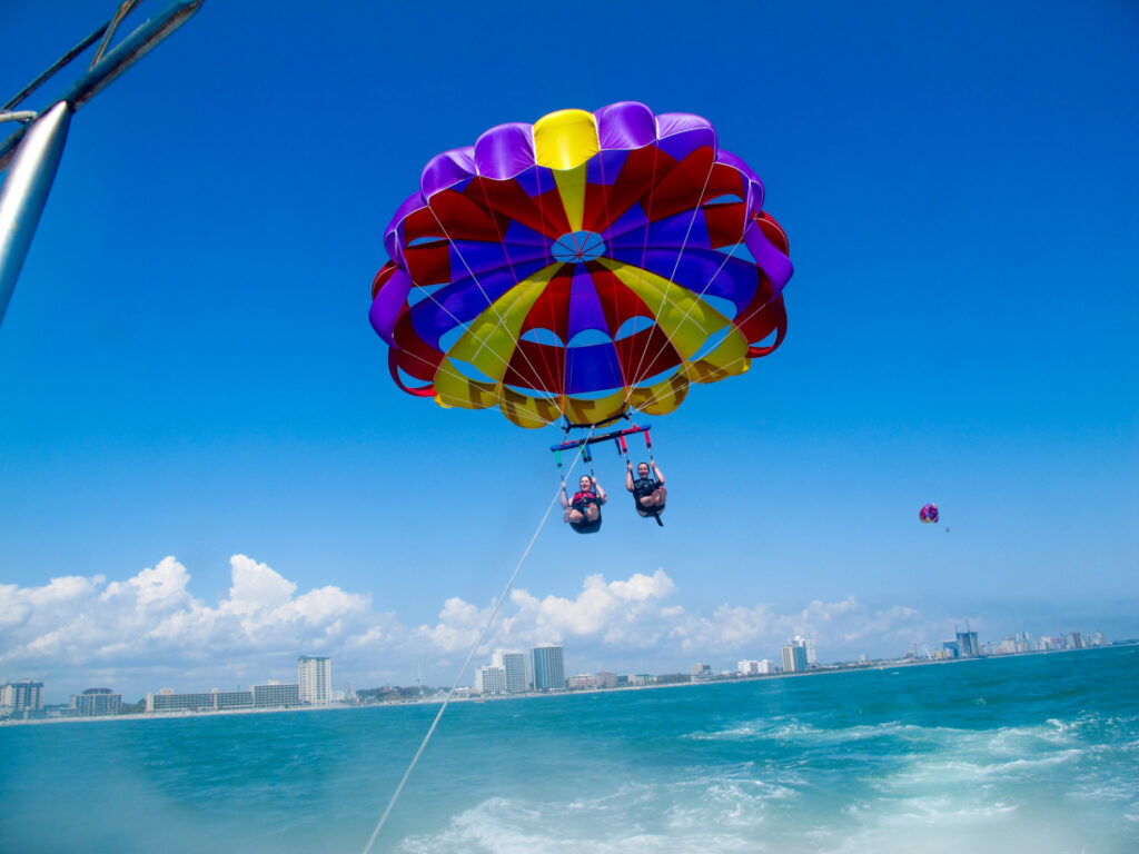 A blue, red, and yellow parasail is open, carrying two people above aqua-colored water on a clear, sunny day. 