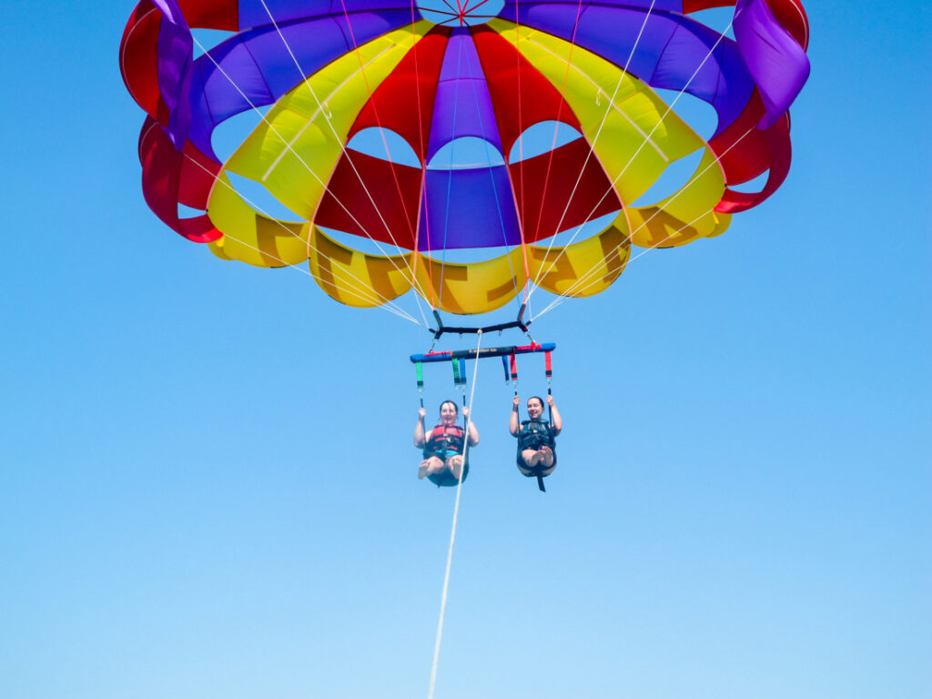 Two parasail riders are in a harness, flying in the sky. Their blue, yellow, and red parasail is open above them, and a rope tows them from below. 