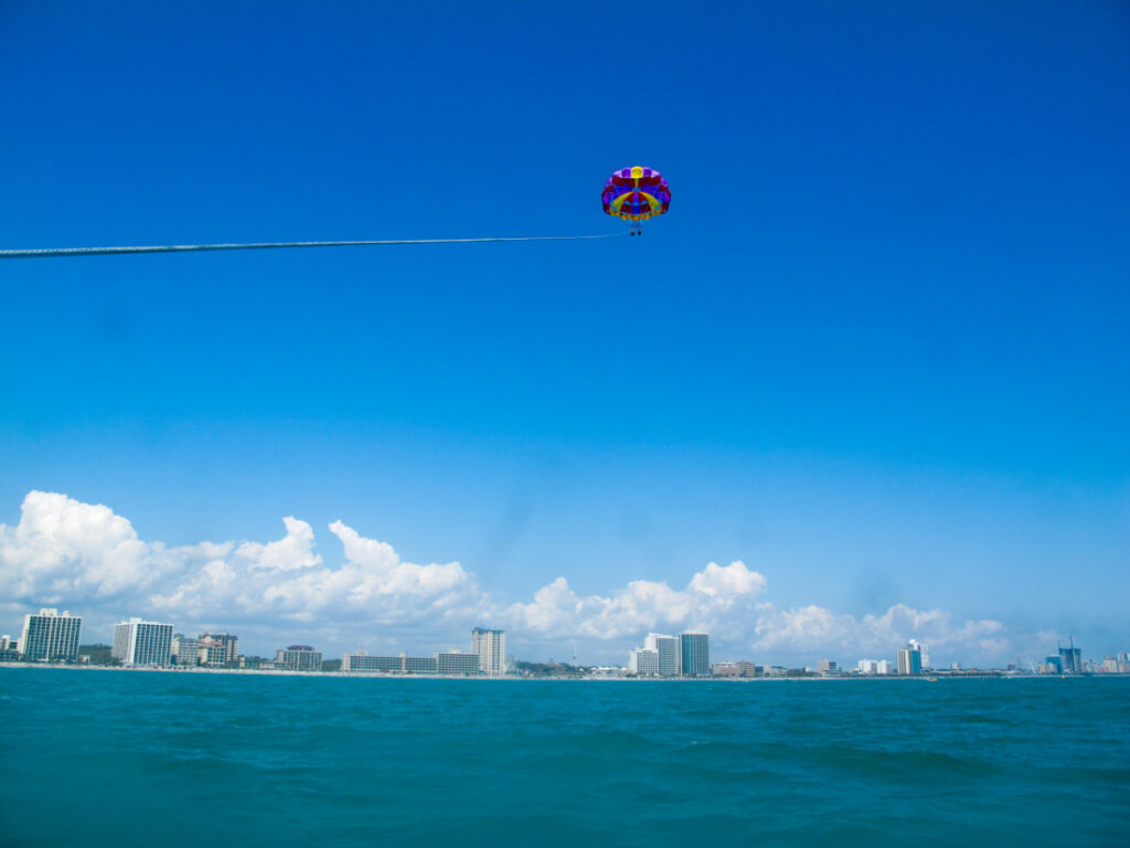 A parasailing flight is shown sailing at a higher altitude against a blue sky. The city skyline is in the background along the horizon.