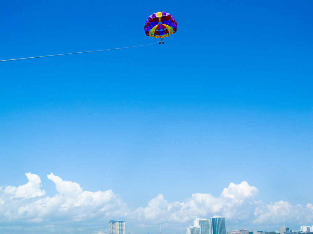 A red, blue, and yellow parasail is open against a blue sky. Along the bottom of the image is the city skyline with a few white fluffy clouds. 