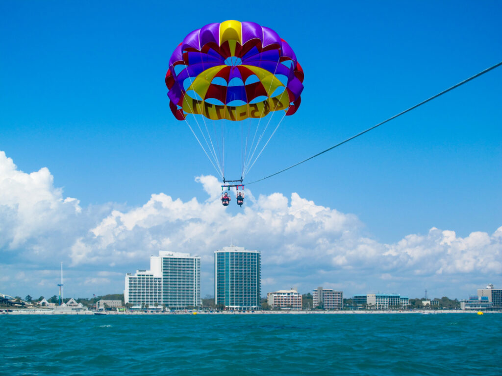 A purple and yellow parasailing canopy is open against a blue sky. Two passengers in the parasailing harness are holding a tow rope as they fly against the backdrop of a city skyline. 