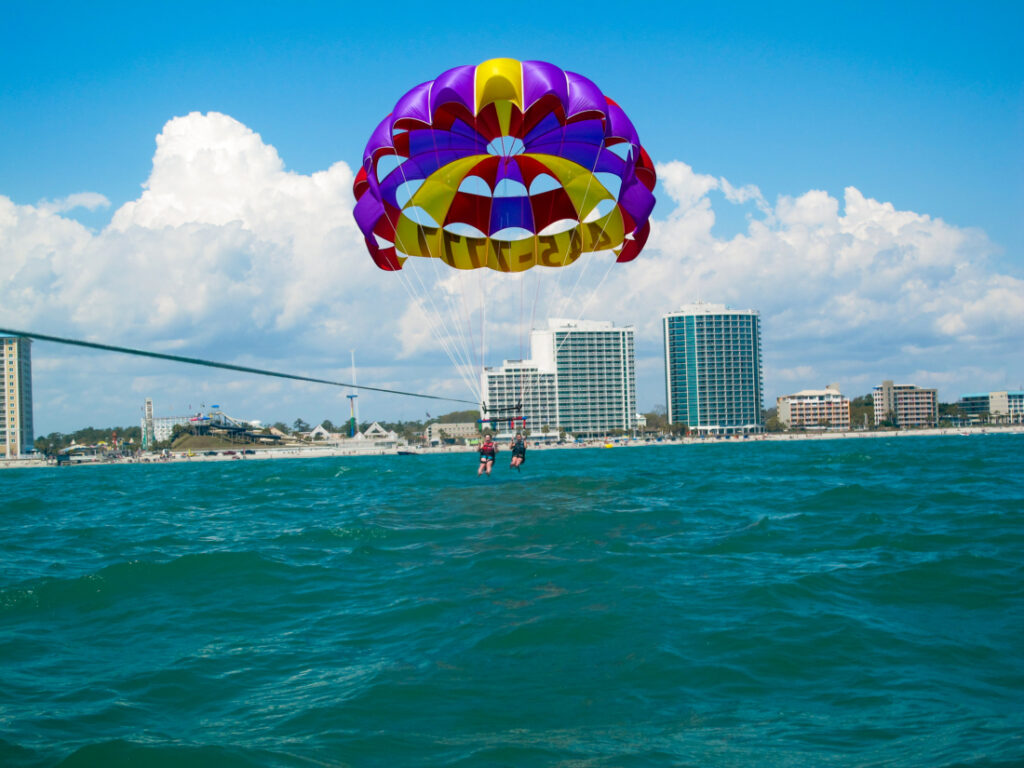 Two parasail riders are coming in for a landing in aqua-colored water. Their parachute is blue, yellow, and red, fully open above them. In the background is a city skyline with a beach. 