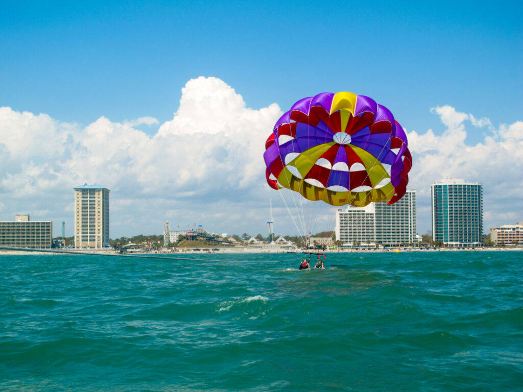 A purple parasailing canopy glides close to the waterline. The riders are getting dipped into the deep turquoise water, with a city skyline in the background. 