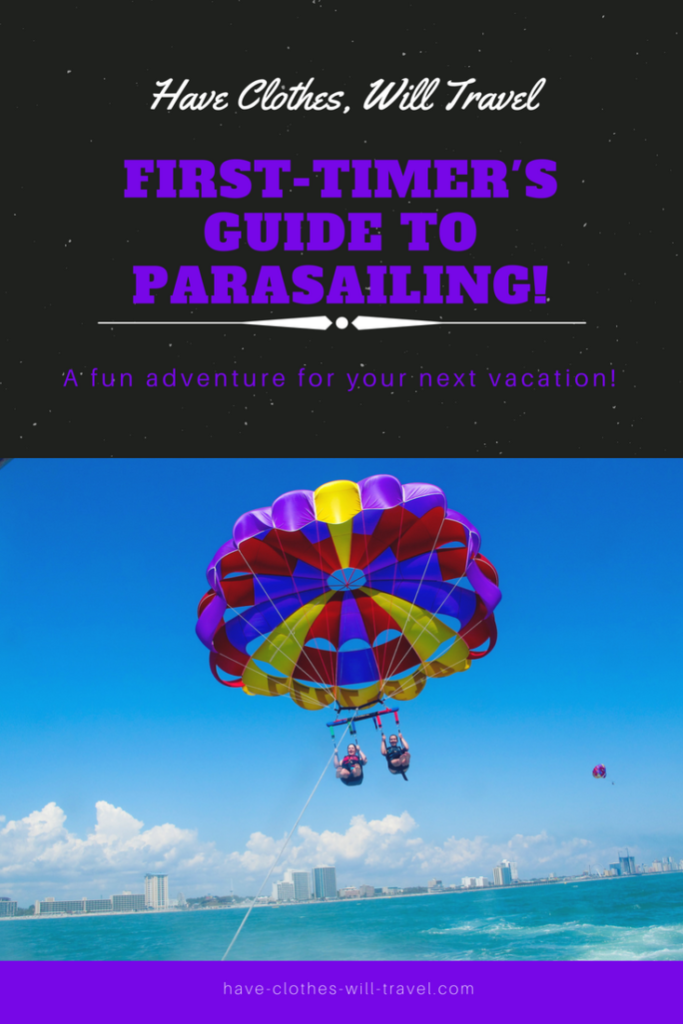 A hero image reads: Have Clothes, Will Travel: First-Timer’s Guide to Parasailing! A fun adventure for your next vacation! The Text is in purple on a black background. The image shows two parasailers riding in a blue, red, and yellow parasail, flying above the water with a city skyline in the background. 