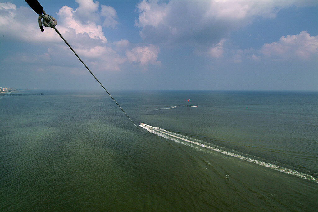 Grey-green water is shown far below the view of a parasailer. A rope tethers the rider to a boat with a white-colored wake trailing behind. 