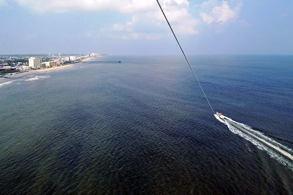 A photo taken while parasailing. The photographer is viewing the rope and boat far below them on the water. In the background, there is a city skyline and blue sky. 