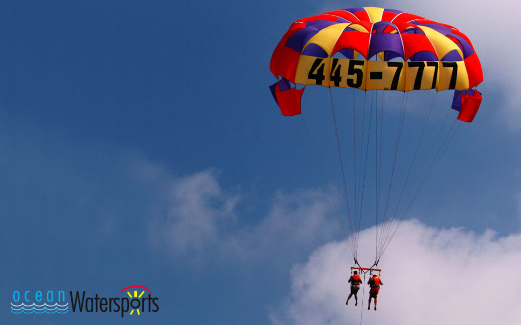 A bright red, blue, and yellow parasail is flying against a blue sky with two riders wearing orange vests. The image reads Ocean Watersports. 
