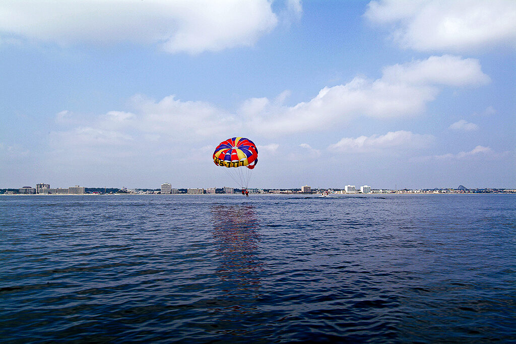 A bright, colorful parasail hovers above the surface of dark blue water. In the background, the sky is sunny and blue with white fluffy clouds. 