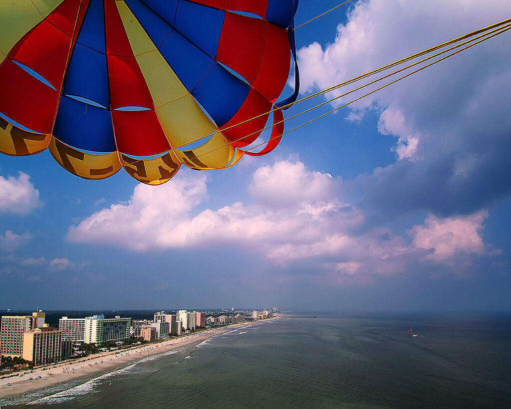 A yellow, blue, and red parasail is open against a blue sky with white fluffy clouds. Beneath the sail is a city beachfront, with a tan sandbar running the length of the grey water. 