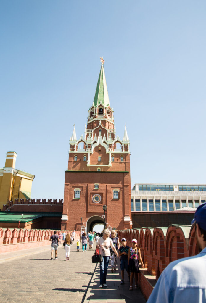 An exterior shot of the Kremlin Armoury building - a tall red brick building with an ornate steeple. Visitors walk on a brick road towards the building, entering the grounds through an archway.