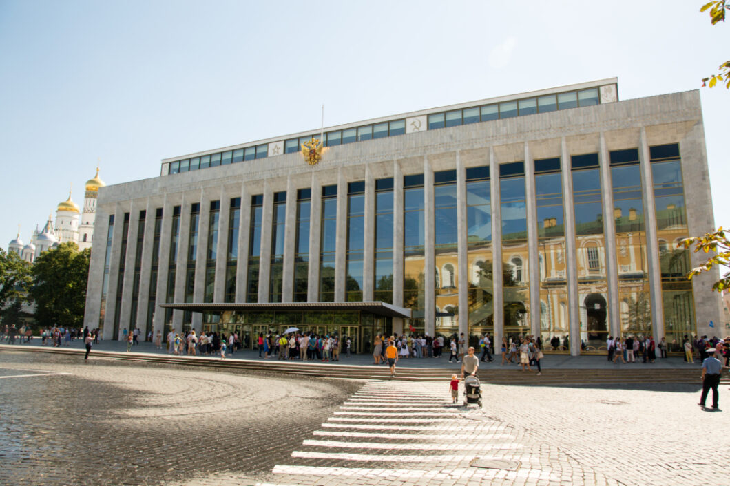 An exterior shot of the front of the State Kremlin Palace Concert Hall - a long steel-gray brick building with floor-to-ceiling glass windows. Visitors gather in front of the building and on the street.