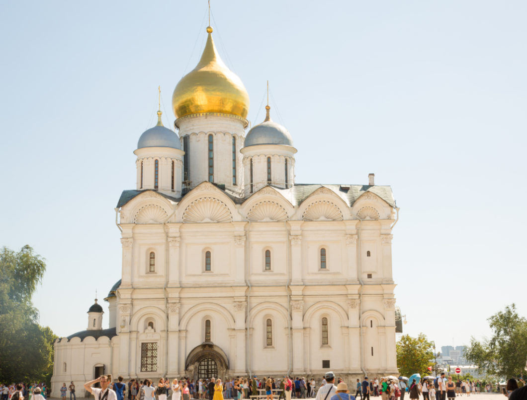 An exterior image of the Archangel Cathedral inside the Kremlin in Russia. The white stone building is several stories tall and boasts three large towers at the top.