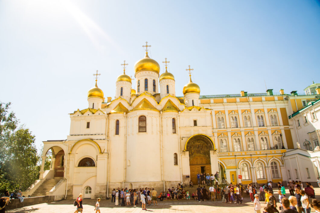 An exterior shot of the The Cathedral of the Annunciation in Russia. The building has a light cream colored exterior with gold trim and gold roofing. People mill about in the courtyard in front of the Cathedral.