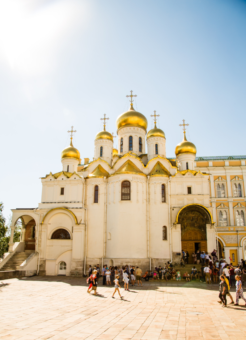 An exterior shot of the Annunciation Cathedral inside the Kremlin. The building is a giant white stone exterior, topped with golden towers. Crowds of visitors walk in and out of the building.