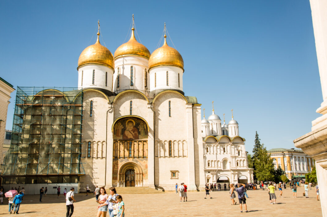 An exterior image of the front of the The Assumption Cathedral inside the Kremlin. The tall building has a white exterior, some of which is covered with scaffolding, and three large bell towers with gold roofs.