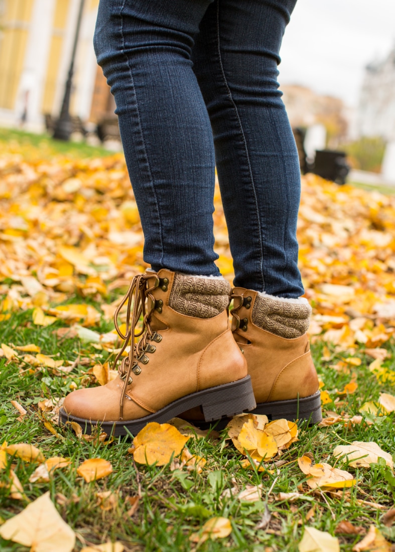 A close up image of a woman's legs. She's wearing blue jeans and tan winter boots. She is standing on green grass covered in golden fallen leaves.