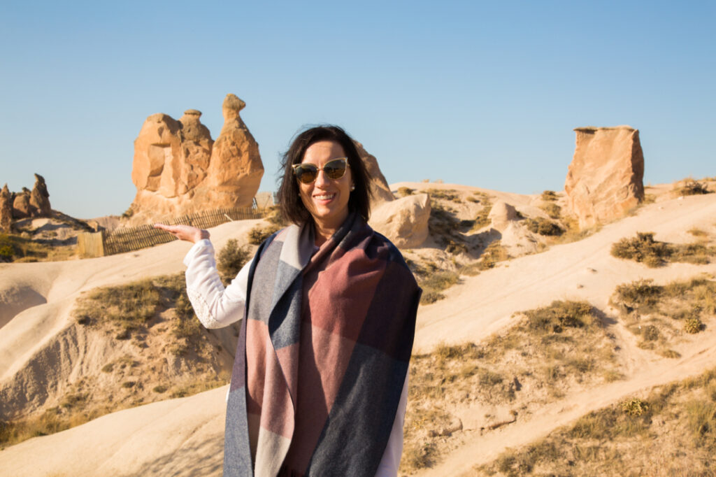 A woman in a scarf standing in front of a rock formation in cappadocia. She appears to be holding a formation that looks like a camel.