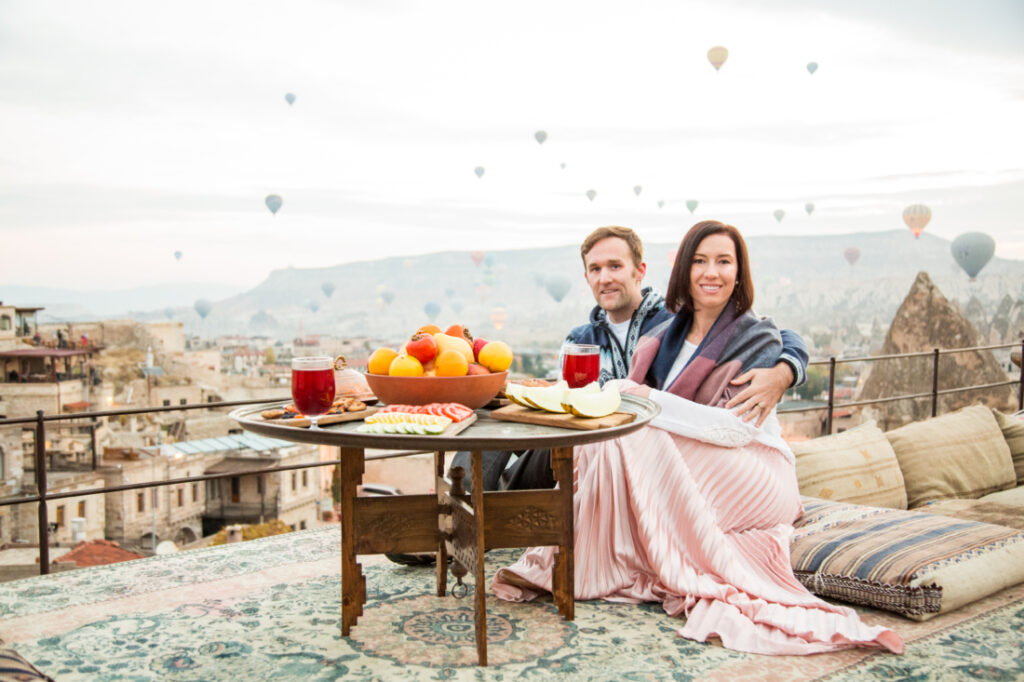 A couple sitting together next to a breakfast table in Cappadocia, as hot air balloons rise behind them