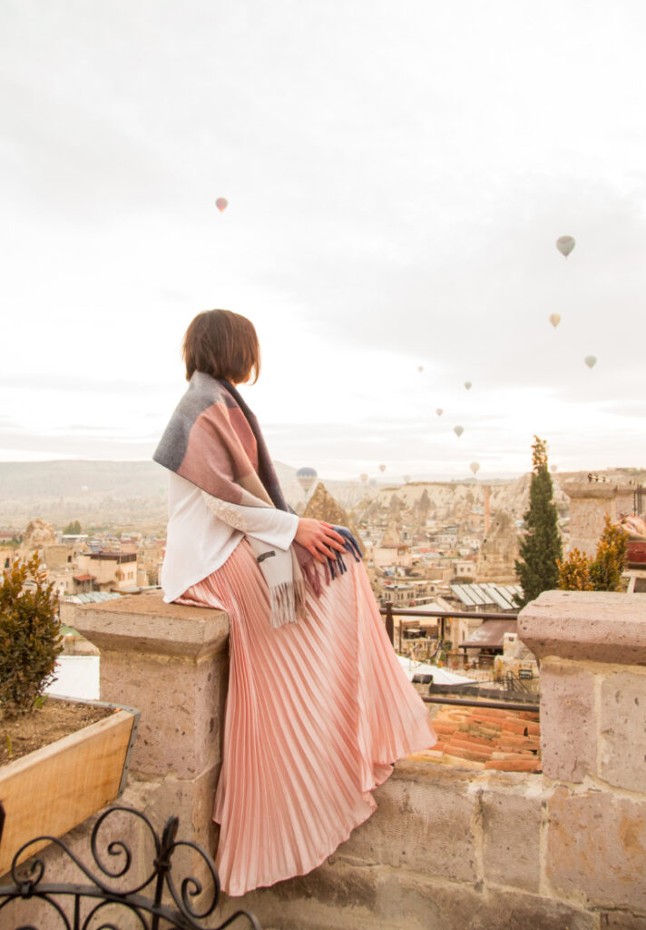 A woman in a pink skirt is sitting on a ledge overlooking a city with hot air balloons.