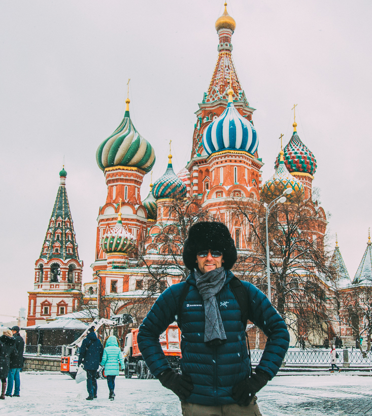 A man poses in front of a colorful Russian building in the middle of a winter scene. He's wearing a navy blue winter jacket and furry black Russian hat, scarf, and gloves.